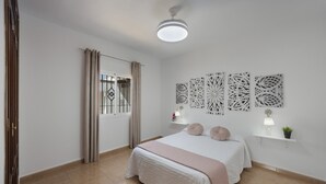 Quarto com cama, ventilador de teto, janela com cortinas, obras de arte na parede e mesa de cabeceira com luminária e planta.