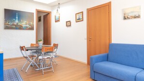A dining area with a table and chairs, a blue sofa, wooden doors, and wall-mounted artwork. - Villa Smeralda 9 – Family Apartment with Pool near Olbia (Telti)
