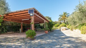 A stone-paved pathway leading to a wooden pergola with a solar panel, surrounded by greenery and a palm tree. - Villa Smeralda 9 – Family Apartment with Pool near Olbia (Telti)