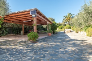 A stone-paved pathway leading to a wooden pergola with a solar panel, surrounded by greenery and a palm tree.