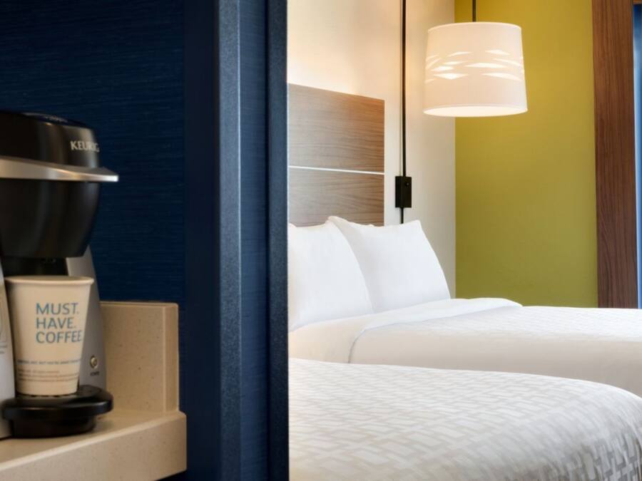 Una habitación de hotel con una cafetera Keurig, una taza que dice 'NECESITO CAFÉ' y una cama con ropa blanca.