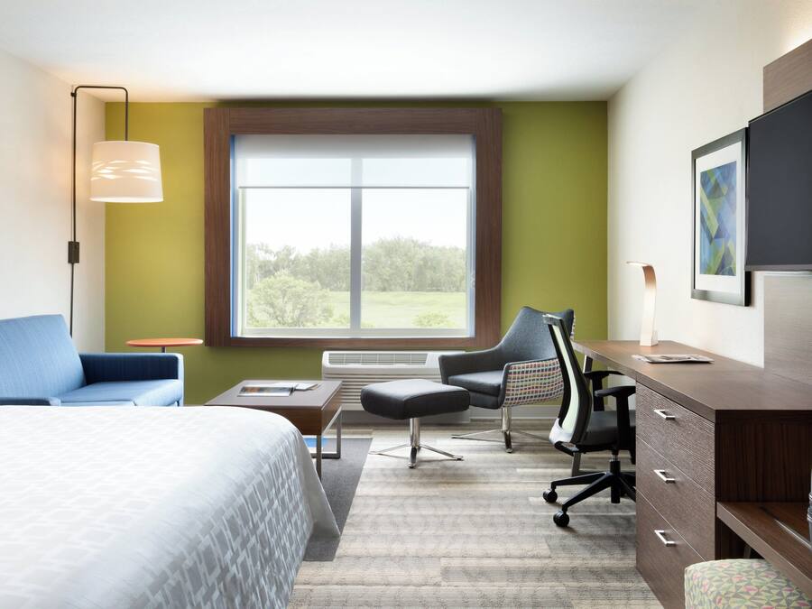 A hotel room with a bed, a desk, a chair, a lamp, and a large window with a view of trees.