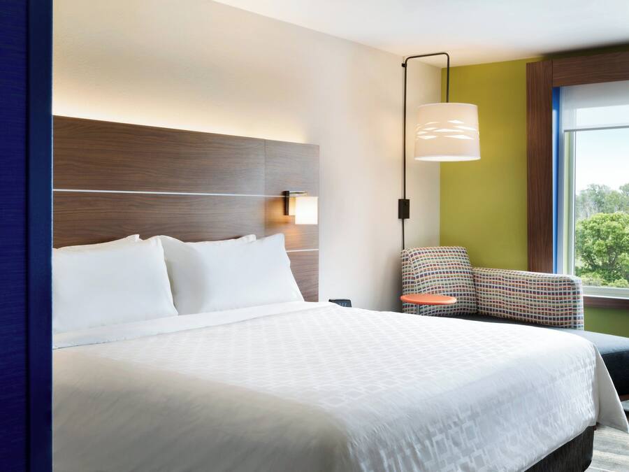 A hotel room with a large bed, a nightstand, a chair, a window with a view of trees, and a wall-mounted lamp.