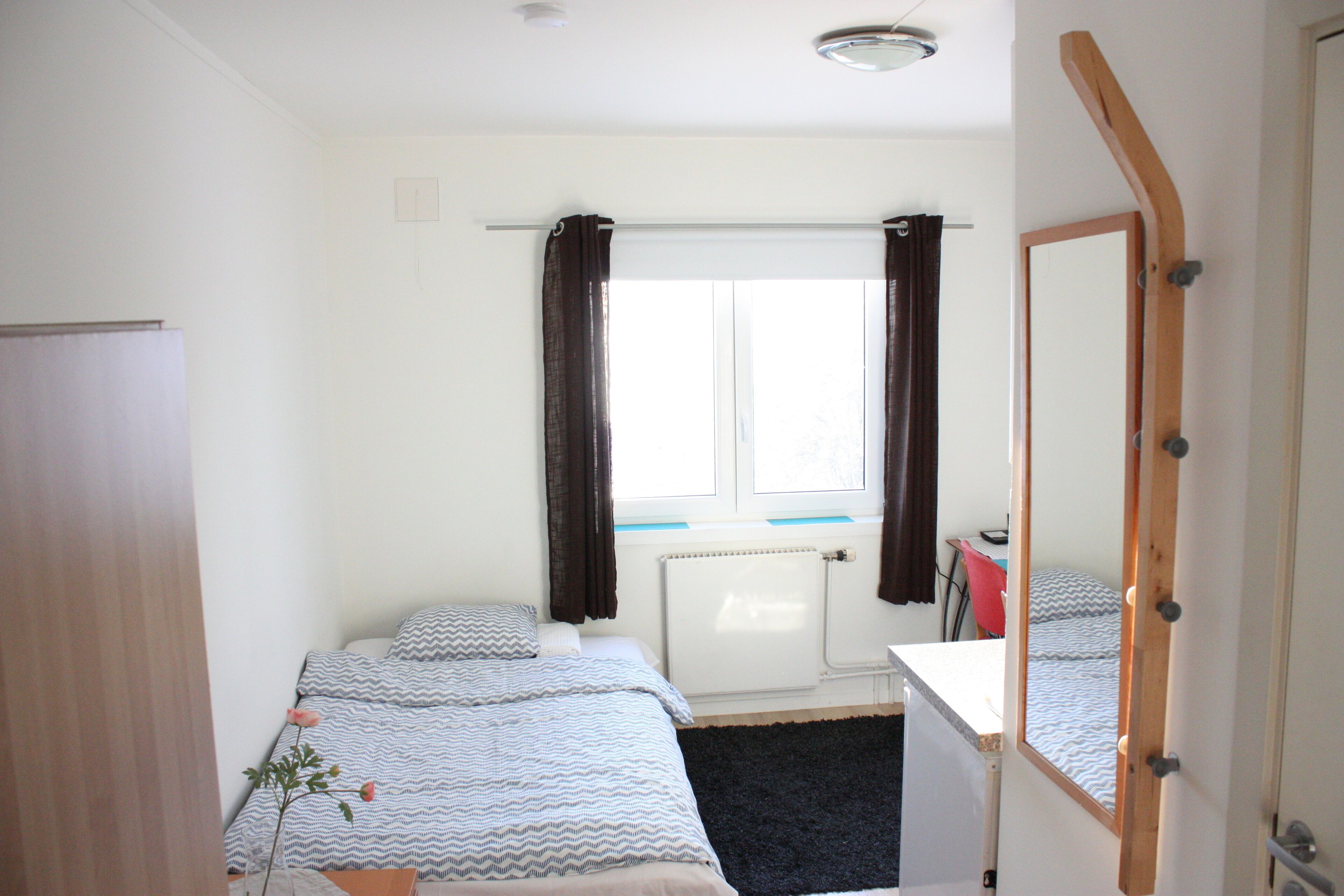 Un dormitorio con una cama, una mesita de noche, un espejo, un radiador y una ventana con cortinas.
