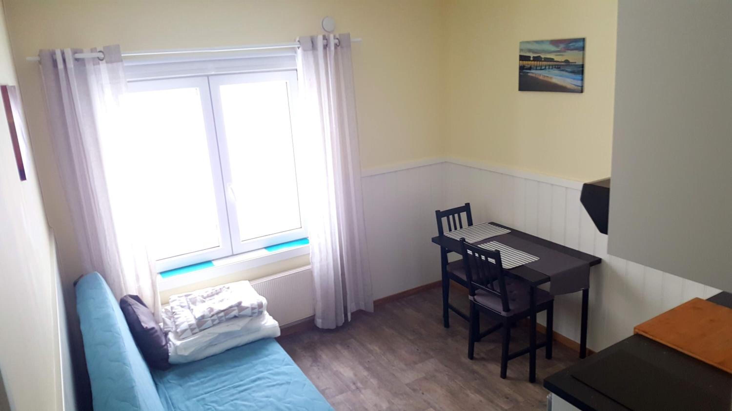 Una habitación pequeña con una cama, una mesa con sillas, un ventanal con cortinas y un cuadro en la pared.