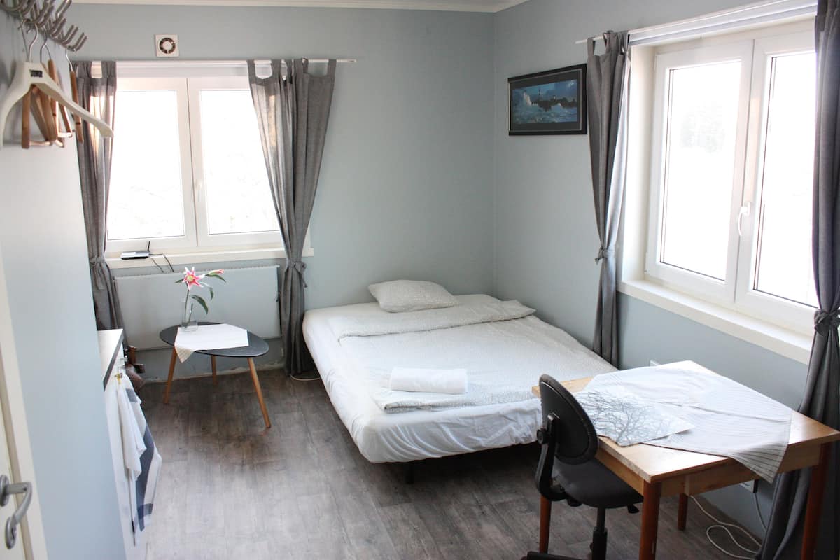 Une chambre à coucher avec un lit, un bureau et une chaise. Il y a une fenêtre avec des rideaux, un tableau au mur et un porte-manteau avec des cintres.