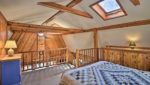 A bedroom with a sloped ceiling, wooden beams, a skylight, a bed with a quilt, a nightstand with a lamp, and a dresser. - Springwater Cabin Near Hiking, Lakes, & Vineyards (Springwater)