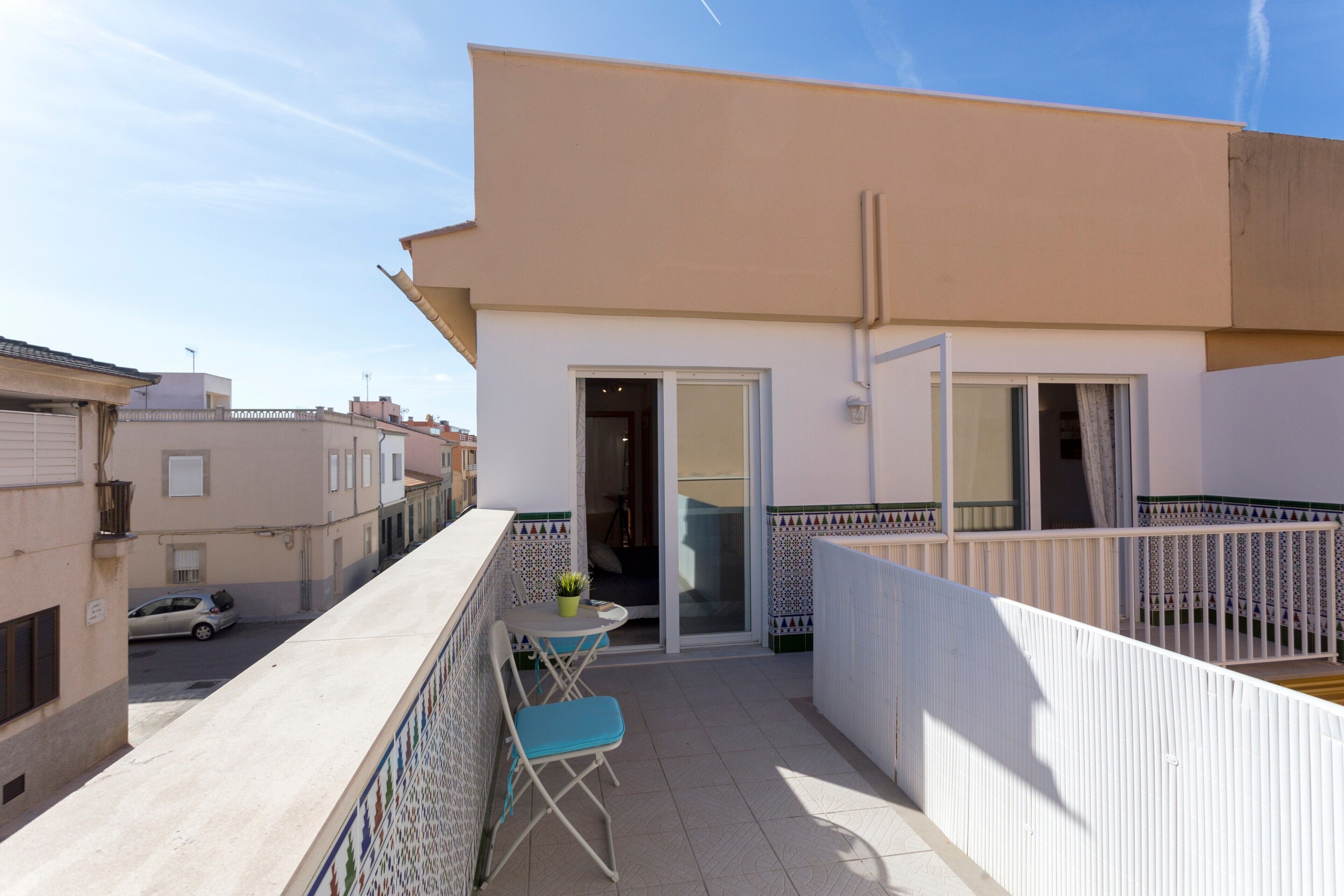 Un balcon avec une table et des chaises, une voiture garée devant, et un immeuble à la façade recouverte de carreaux.