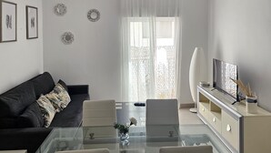 Una sala moderna con una mesa de comedor de vidrio, un sofá negro, un televisor sobre un mueble bajo y decoración en la pared.