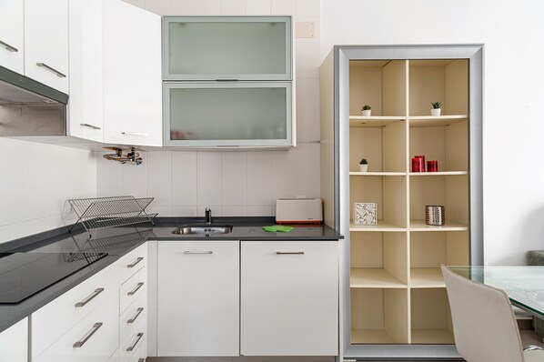 Una cocina moderna con armarios blancos, encimera negra, horno empotrado y despensa con puerta de vidrio y estantes.