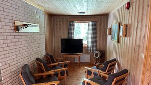 A cozy living room with wooden walls and floors, a television, and four wooden chairs with cushions. - Torpomoen (Al)
