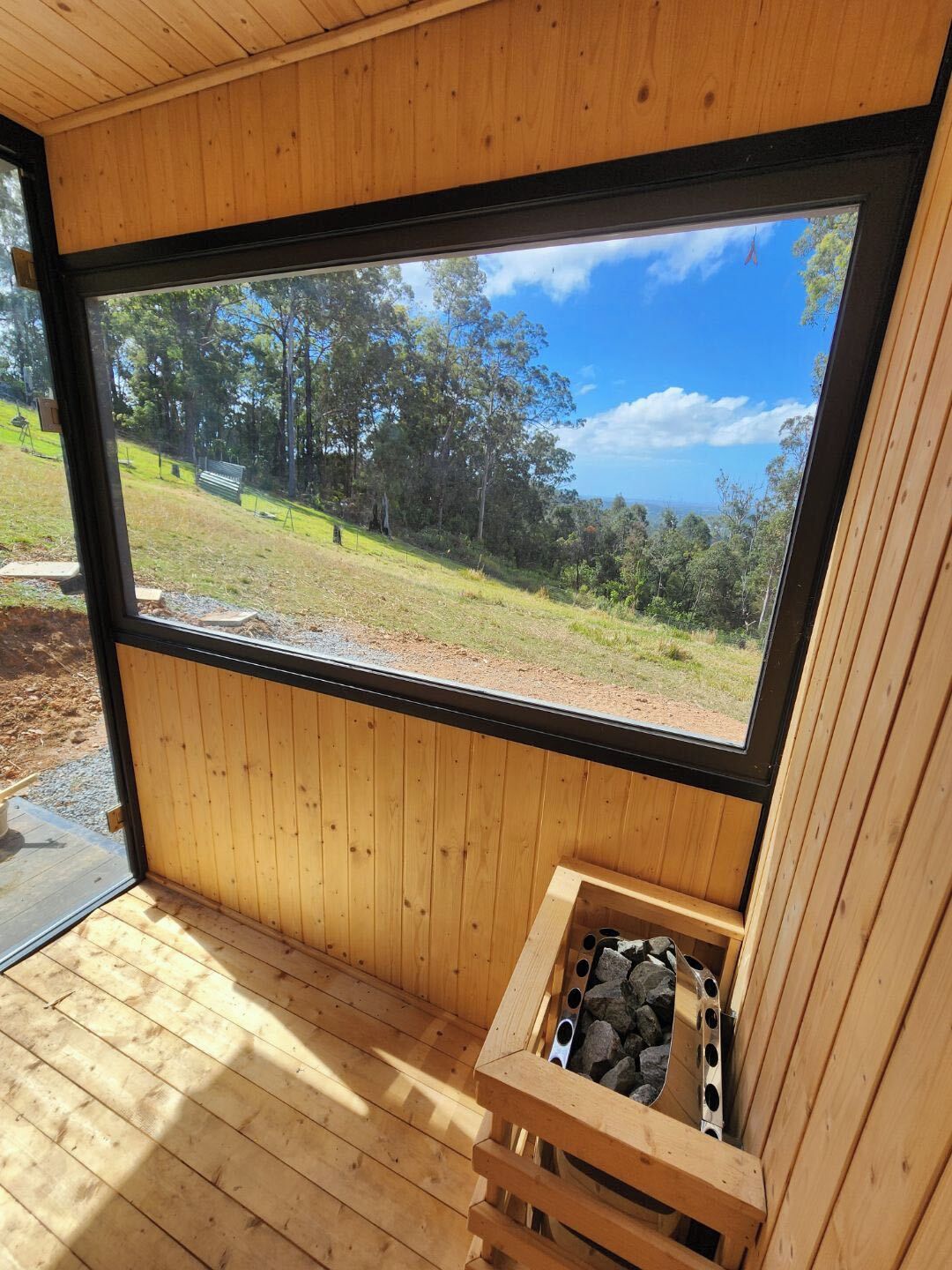Ein Holzinterieur mit einem großen Fenster, eine integrierte Sauna mit Steinofen und Blick auf eine grüne Fläche mit Bäumen.