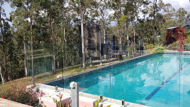 A rectangular swimming pool with clear blue water, surrounded by a glass fence, and set against a backdrop of trees and a building. - Treehouse #3, Tri-Level, Large BBQ. Amazing views (Austinville)