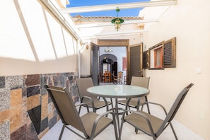 A covered outdoor patio with a round table and chairs, a tiled wall, and a view into a building with wooden shutters.