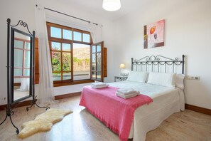 Un dormitorio con cama, ventana con cortinas, espejo y un cuadro en la pared.