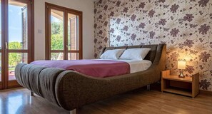A bedroom with a bed, a nightstand, and a window with blinds. - Junior Villa AZ02 - Lania (Kouklia)