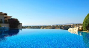 A clear blue infinity pool with a view of a hillside and distant buildings. - Junior Villa AZ02 - Lania (Kouklia)