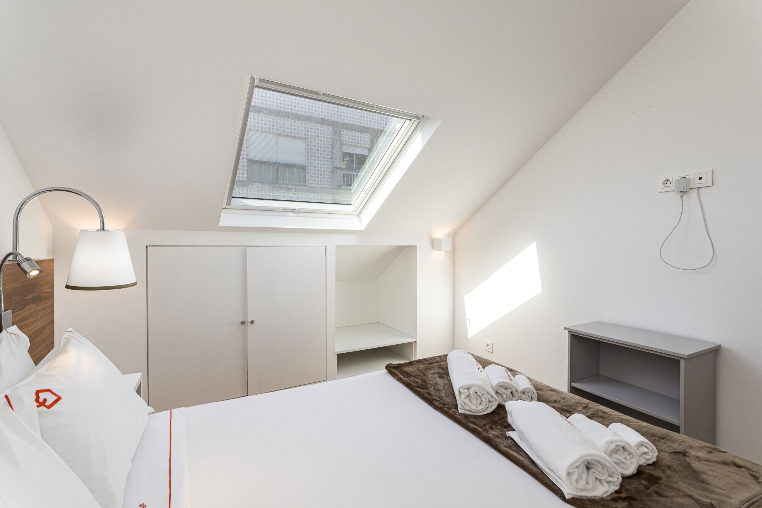 A hotel room with a skylight, a bed with white linens, a bedside lamp, a small desk, and a towel rack.