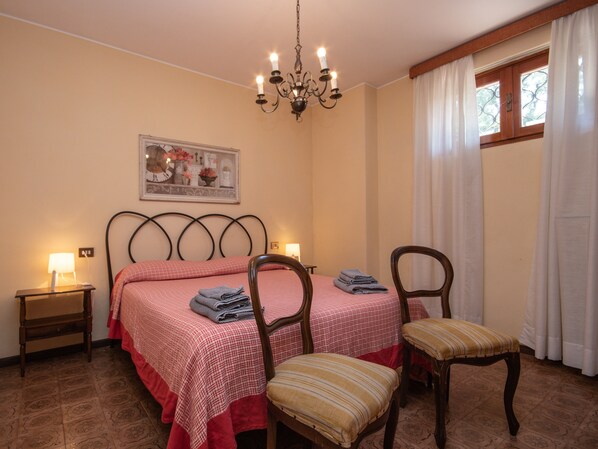 Une chambre à coucher comprenant un lit, deux chaises, une table de chevet, une lampe, un lustre, une fenêtre avec des rideaux et un tableau encadré accroché au mur.