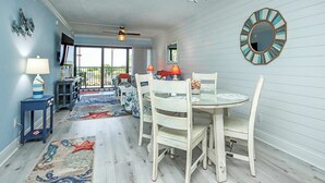 Uma área de refeições com uma mesa redonda e cadeiras, um ventilador de teto e vista para o exterior através de uma porta de vidro.