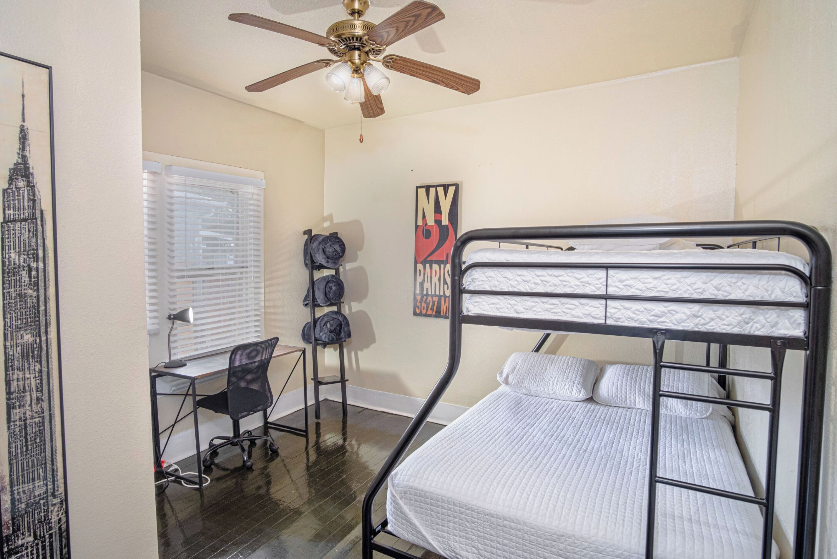 A dorm-style room with a bunk bed, a desk with a chair, a ceiling fan, and a window with blinds.