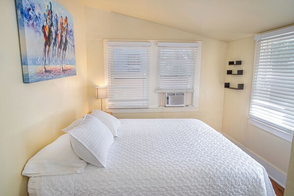 A bedroom with a bed, white bedding, an air conditioning unit, and a painting on the wall.