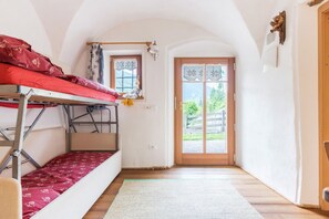 Ein Zimmer mit einem Etagenbett, einem Fenster mit Spitzenvorhängen, einer Holztür und Blick auf Grünfläche.