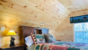 Une chambre confortable dans un chalet, avec un plafond en bois, un lit recouvert d’une courtepointe aux couleurs vives, une table de chevet avec une lampe et une fenêtre donnant sur la forêt.