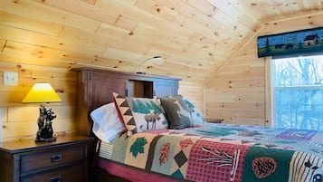 A cozy cabin room with a wooden ceiling, a bed with a colorful quilt, a nightstand with a lamp, and a window with a view of trees.