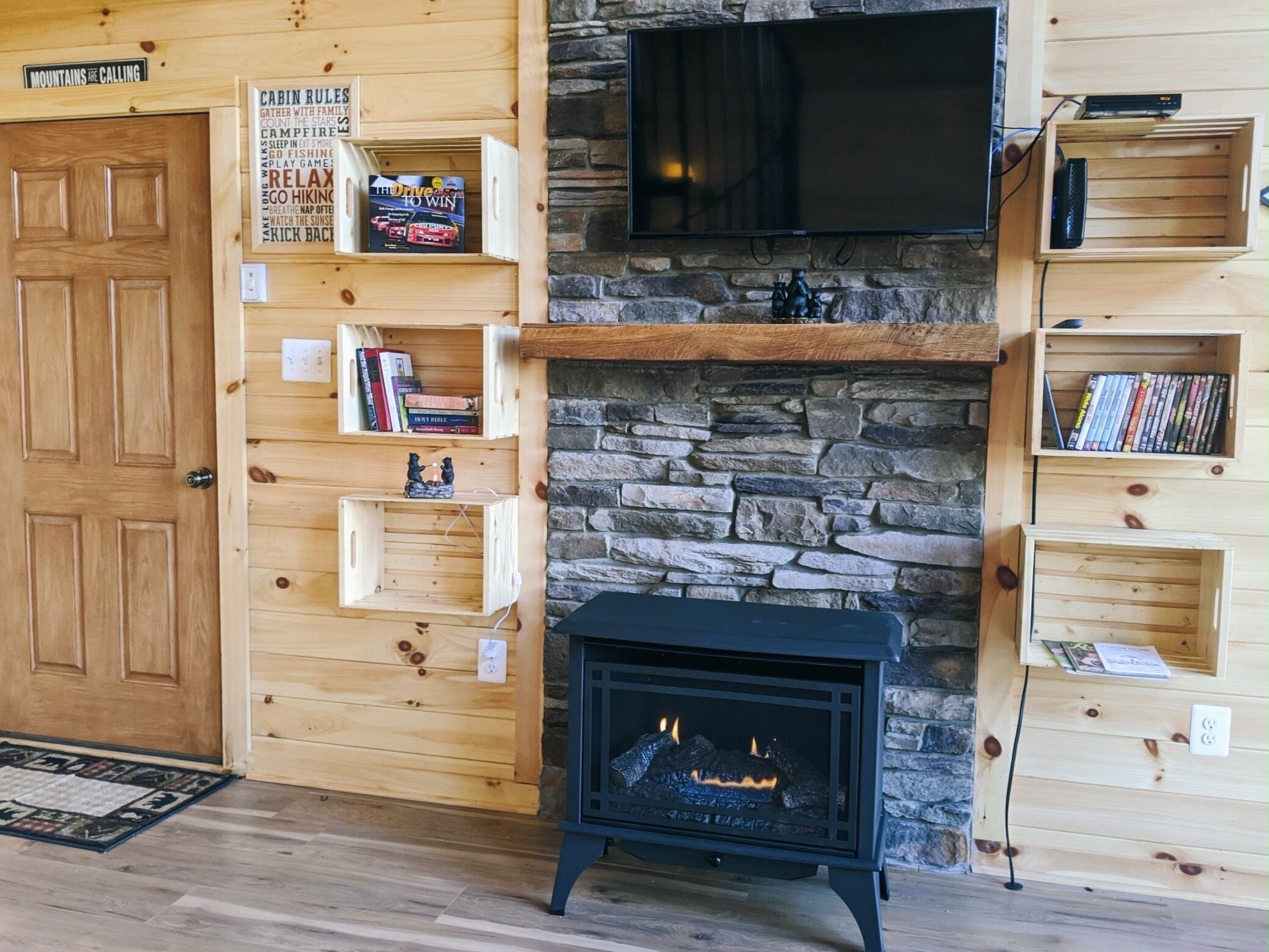 A cozy cabin interior with a stone fireplace, wooden shelves with books and decorations, and a television mounted above the fireplace.