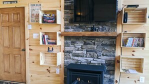 L’intérieur chaleureux d’un chalet avec une cheminée en pierre, des étagères en bois ornées de livres et de décorations, et un téléviseur fixé au-dessus de la cheminée.