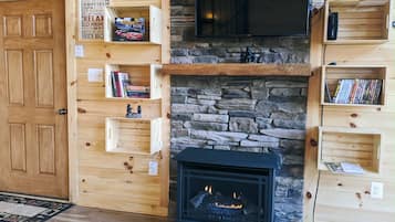 A cozy cabin interior with a stone fireplace, wooden shelves with books and decorations, and a television mounted above the fireplace.