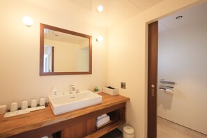 Une salle de bain avec un meuble-lavabo en bois, un lavabo blanc et un miroir au-dessus. On y trouve des articles de toilette sur le comptoir et une poubelle dans un coin.