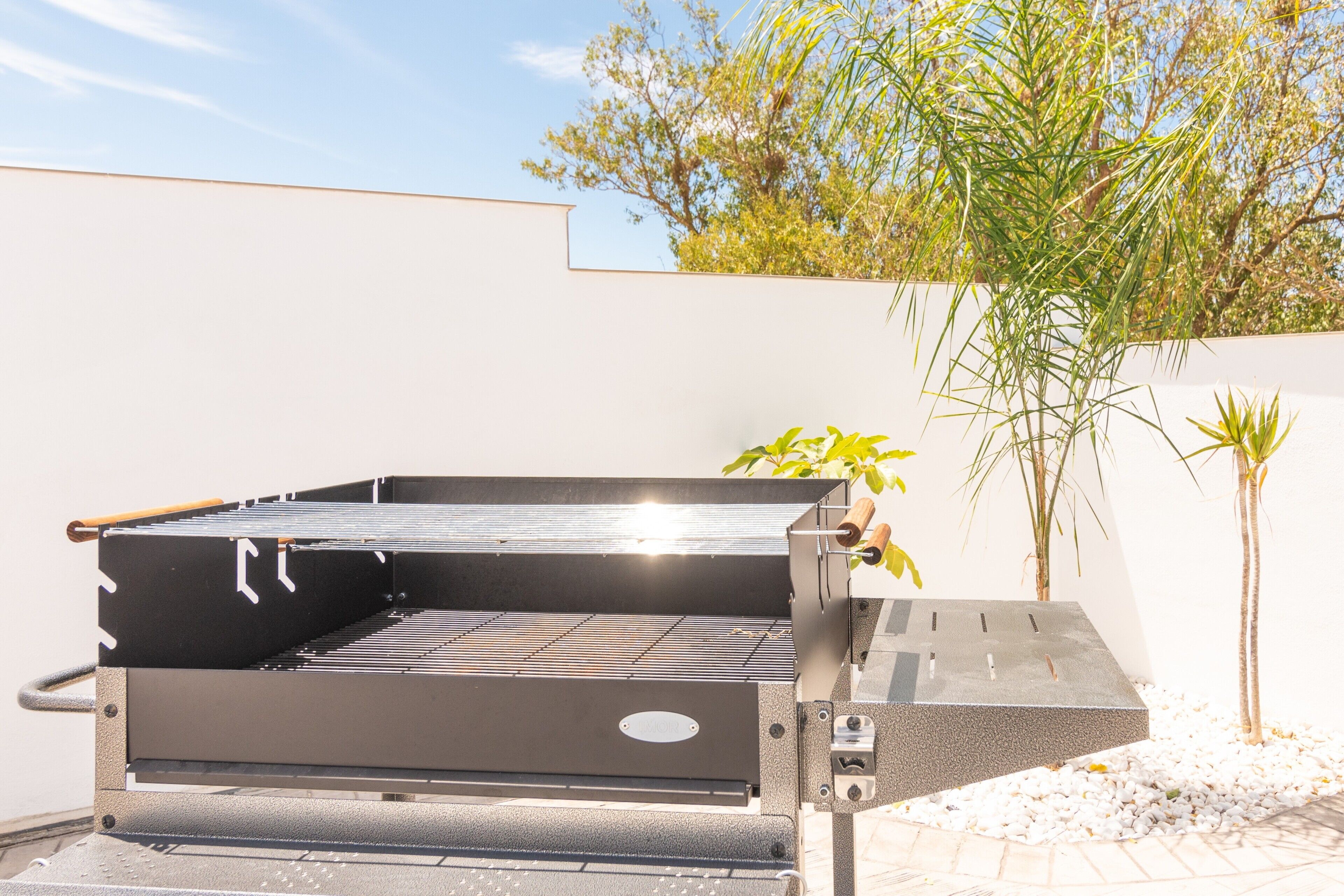 A modern outdoor grill with a flat top and side ventilation, set against a white wall with a tree and a small palm plant nearby.