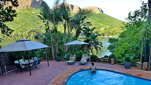A pool area with a view of a lake and mountains, surrounded by lush greenery and outdoor seating. - The Shore House (Hartbeespoort)