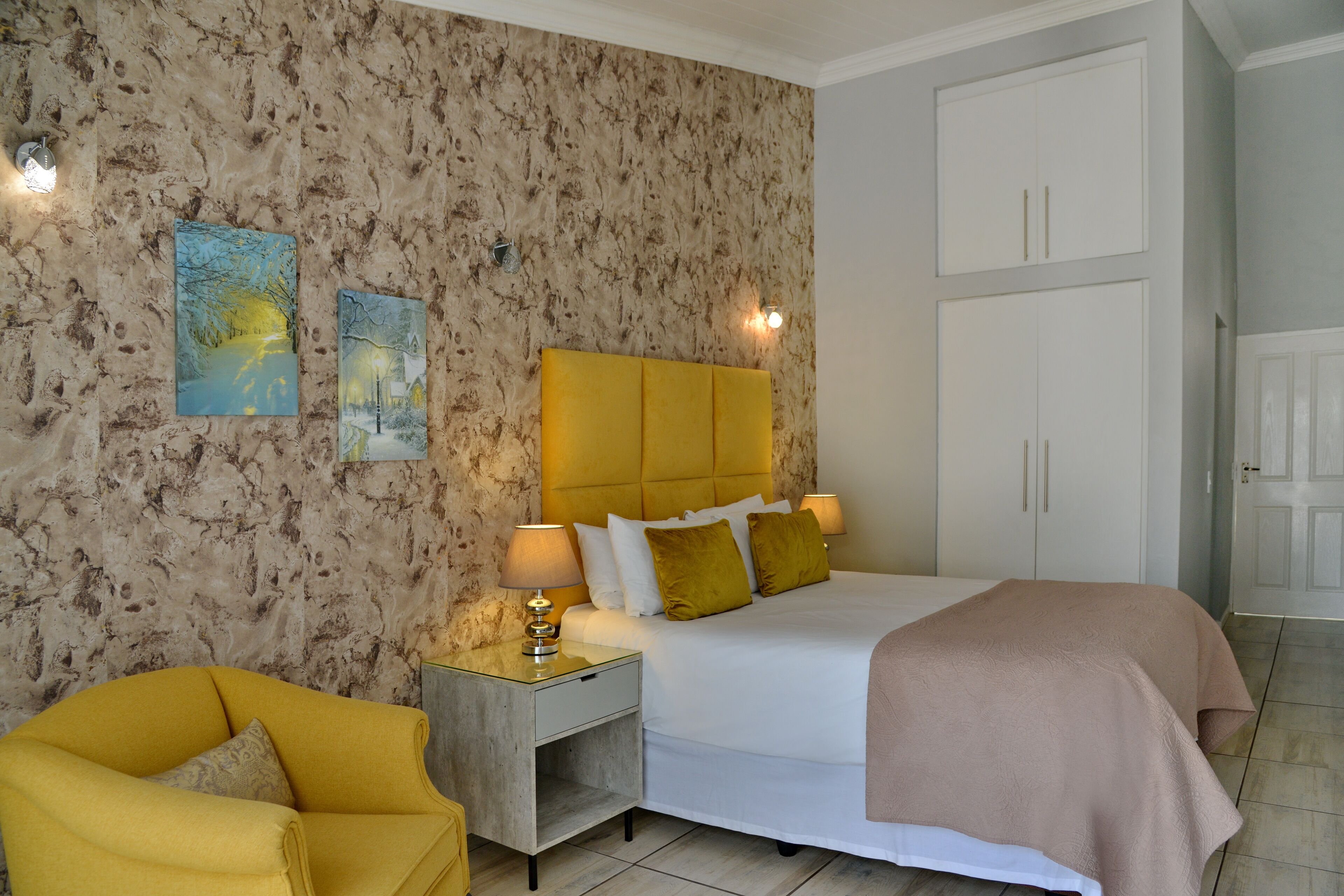 Een slaapkamer met een wand met marmerlook, een bed met een geel hoofdeinde, twee bedlampen, een nachtkastje en een gele fauteuil.