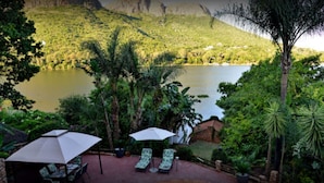 A patio area with lounge chairs and umbrellas overlooking a lake, surrounded by lush greenery and a hilly landscape. - The Shore House (Hartbeespoort)