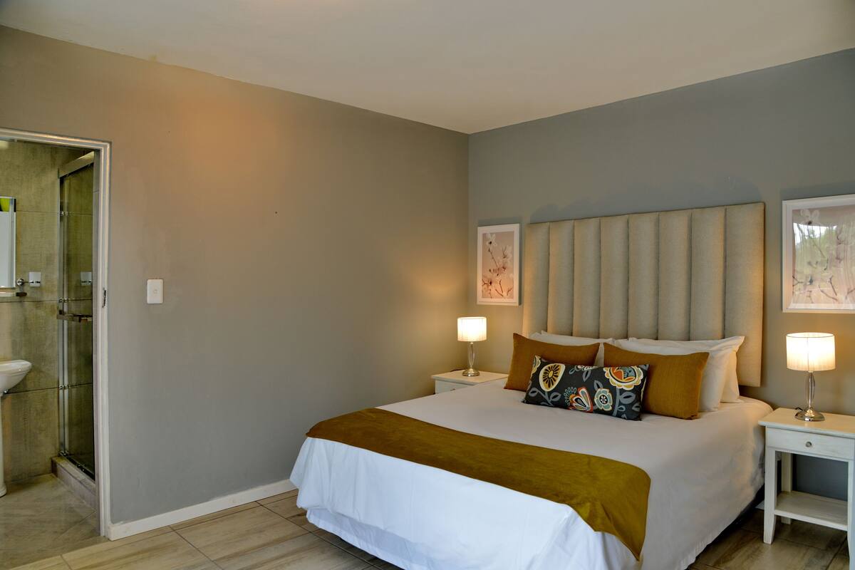 A bedroom with a bed, nightstands, a bedside lamp, a headboard, and a bathroom with a shower and toilet.