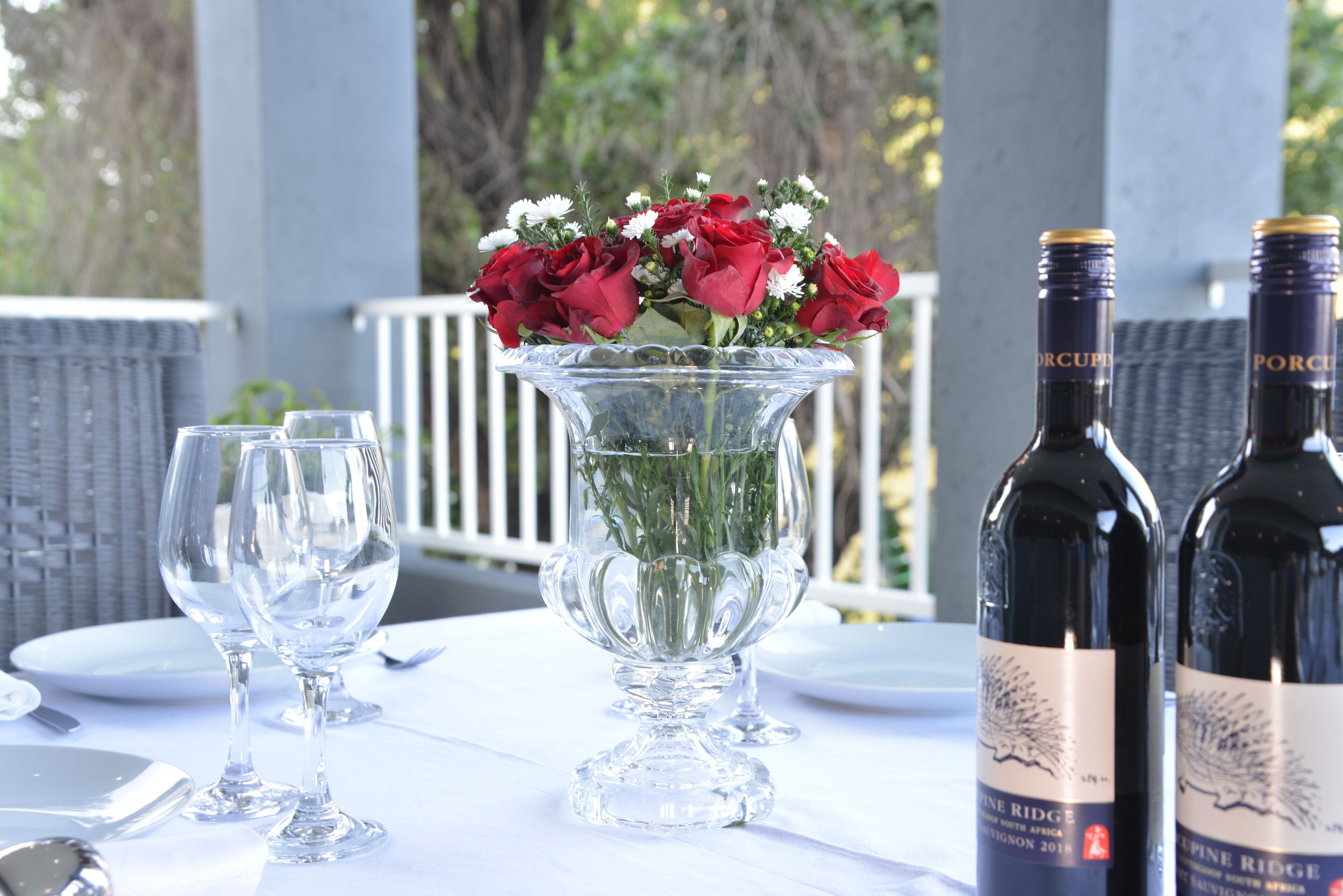 Une table dressée avec des verres à vin, un vase rempli de fleurs rouges et deux bouteilles de vin.