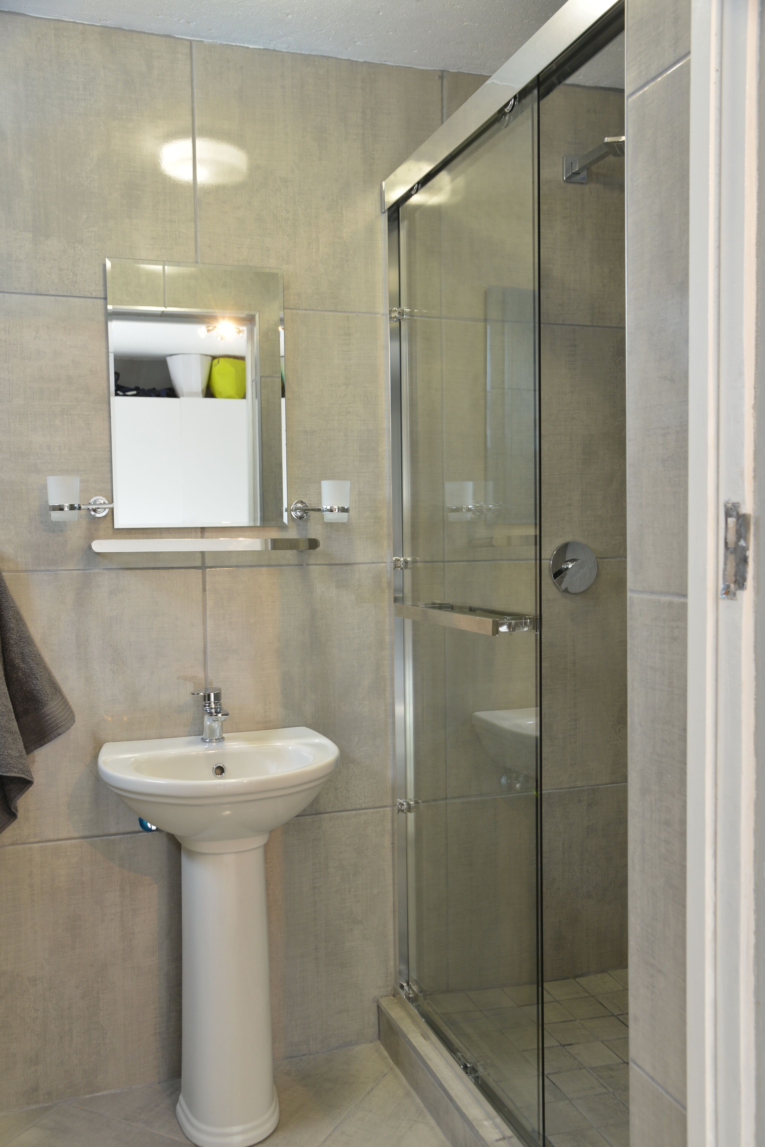 Un baño con lavamanos pedestal, cabina de ducha con mampara de vidrio y espejo sobre el lavamanos.