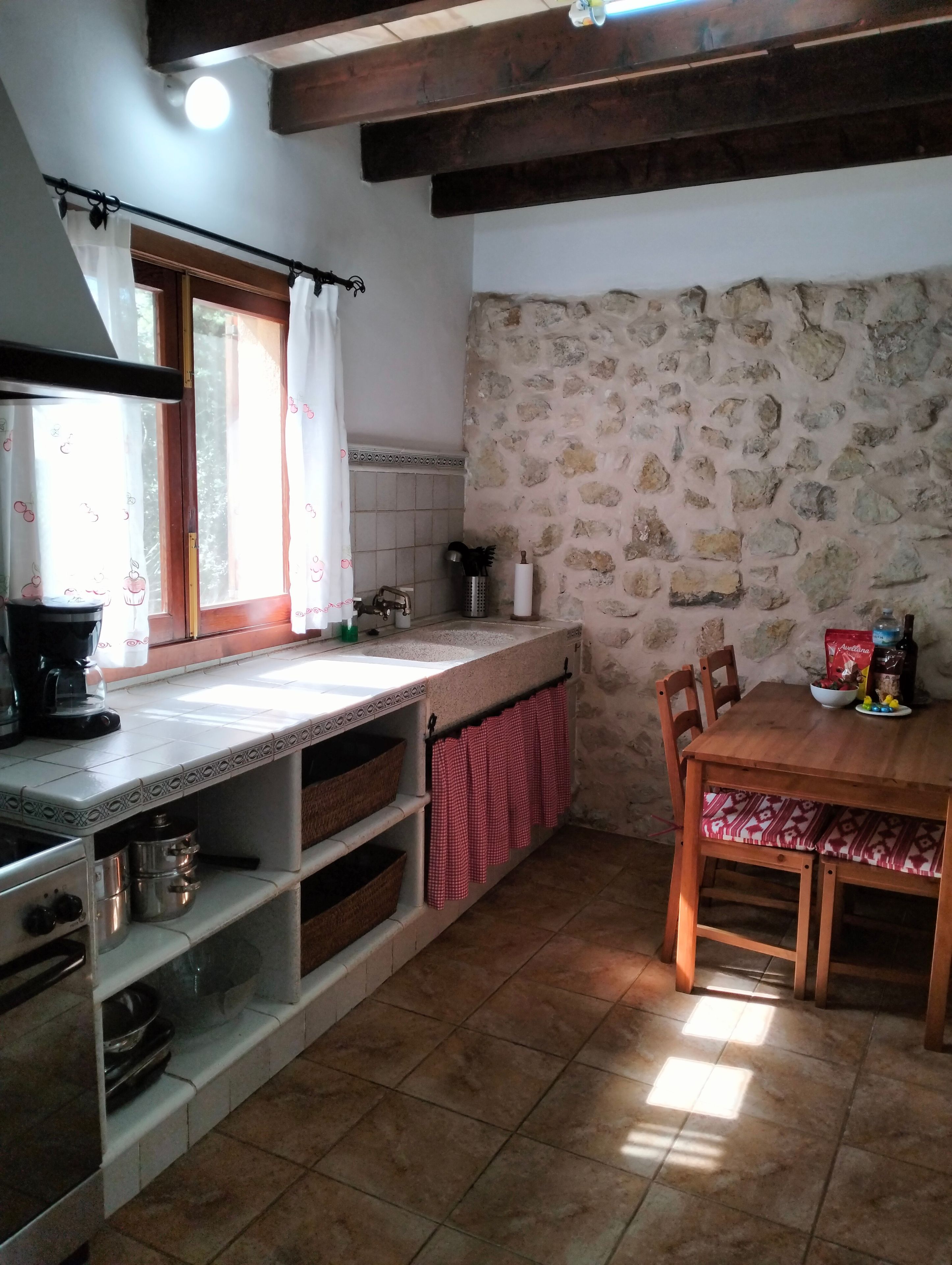 Une cuisine avec un mur en pierre, des poutres en bois, un sol carrelé et une table à manger avec des chaises.
