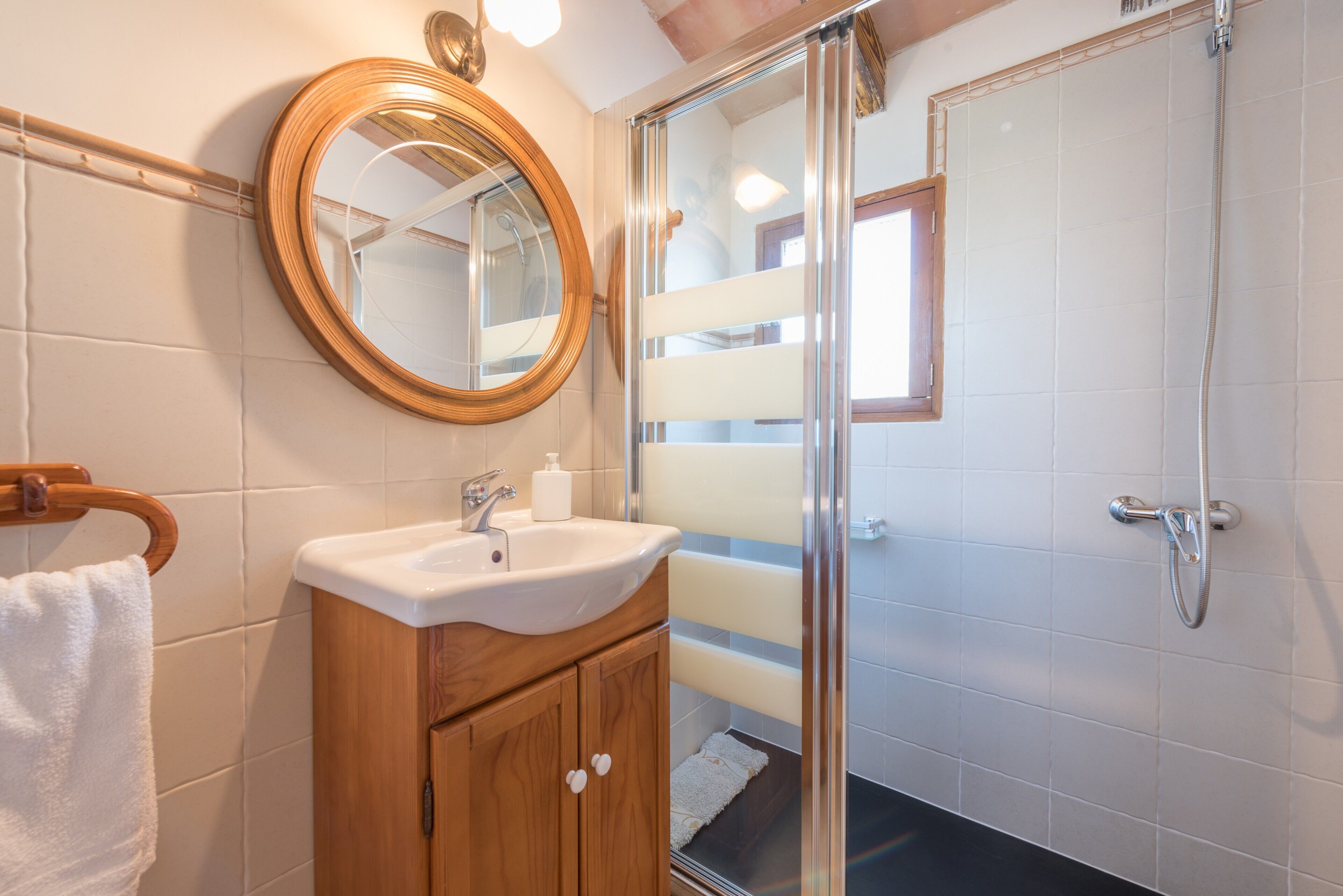 A bathroom with a wooden vanity, a round mirror, a glass shower enclosure, and a white sink.