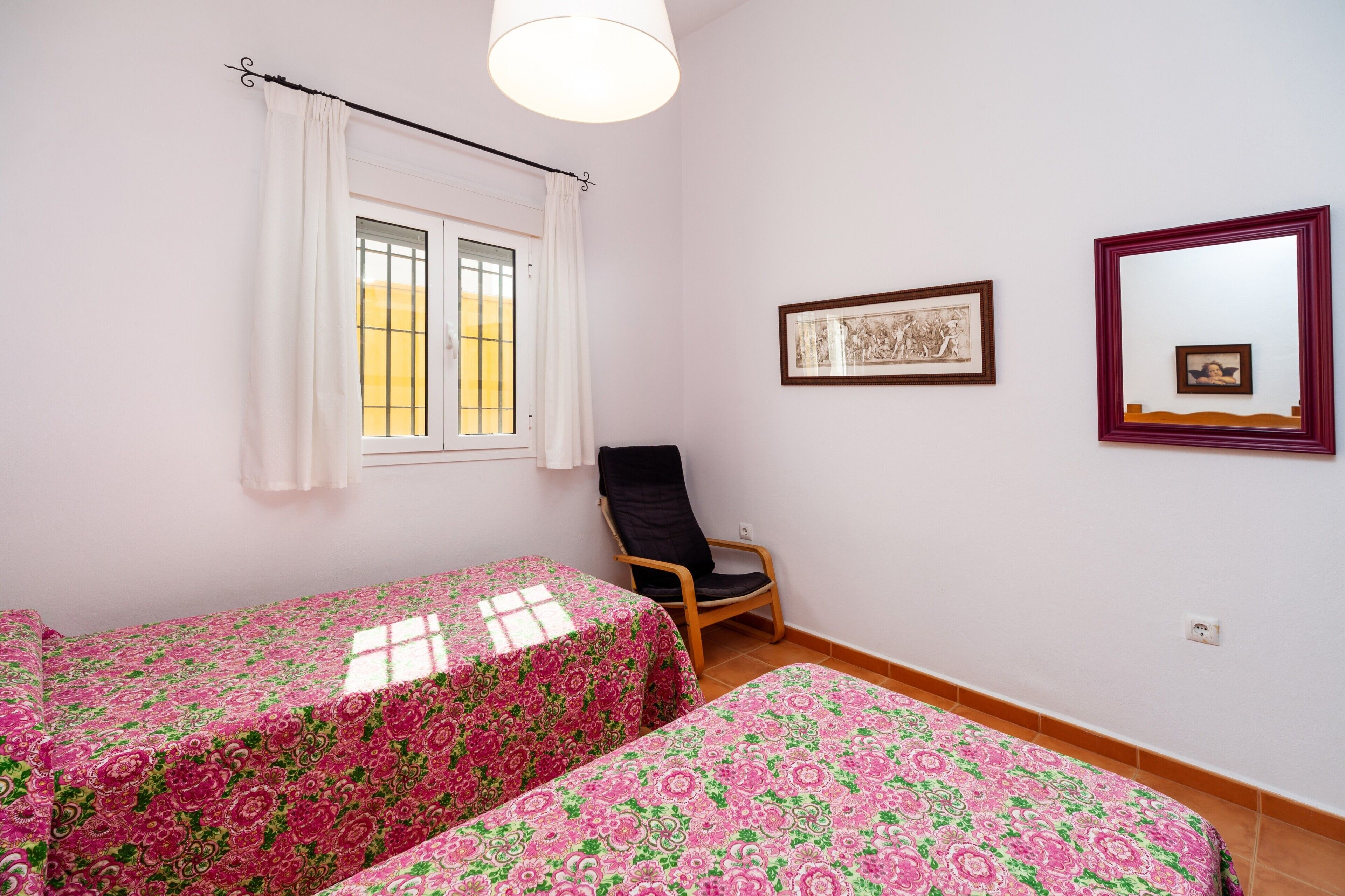 Una habitación con dos camas, una silla, una ventana con cortinas y cuadros en la pared.