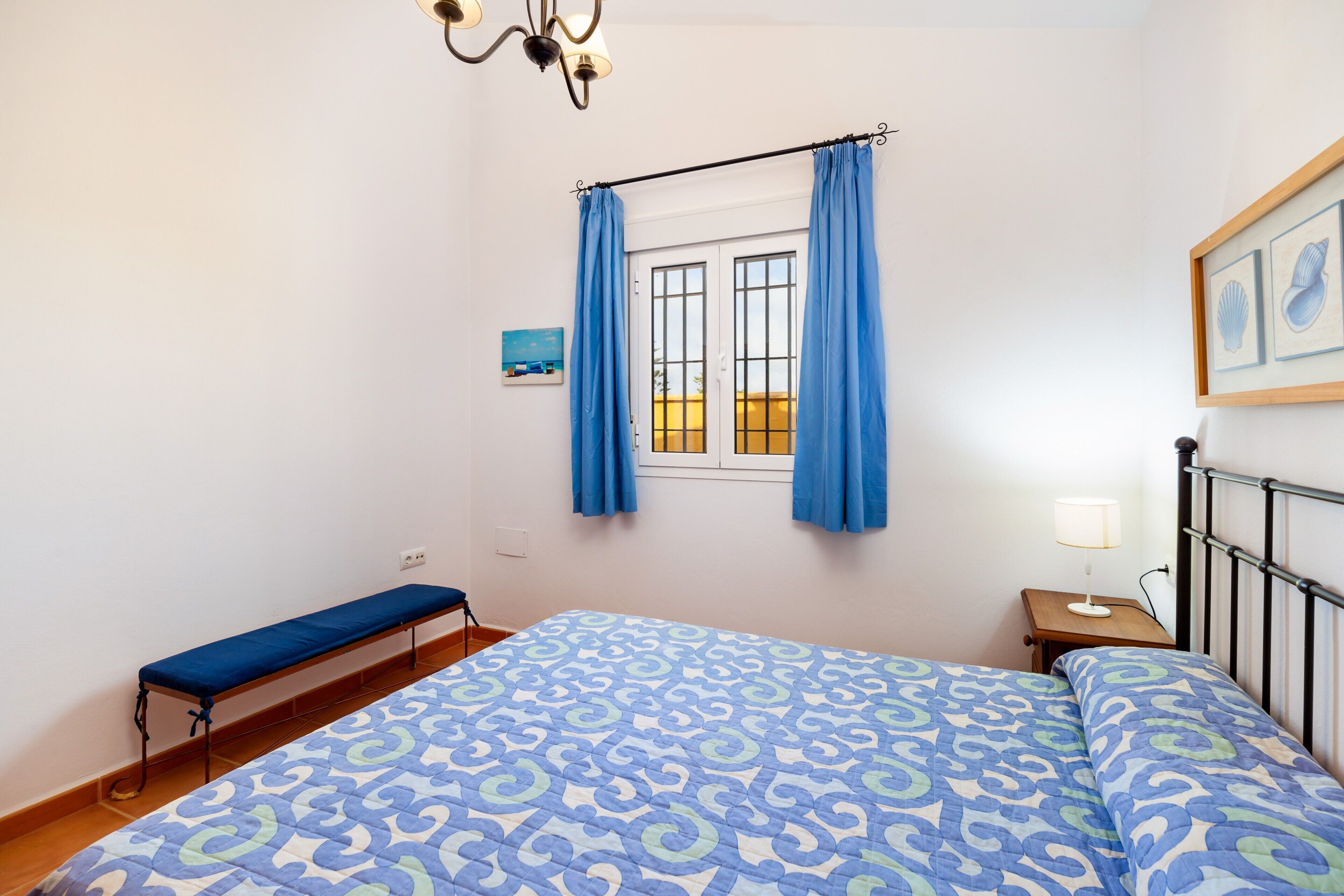 Quarto com cama, um banco, janela com cortinas, mesa de cabeceira com abajur e um quadro na parede.