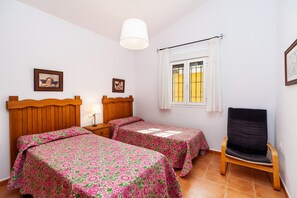 Un dormitorio con dos camas, cabeceros de madera, una mesita de noche, una silla, un ventanal con cortinas e imágenes enmarcadas en la pared.