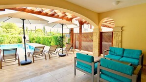Un patio iluminado por el sol con piscina, mobiliario exterior y vistas a la vegetación.