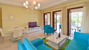 Una sala de estar moderna con televisor, sofás azules, un comedor y un balcón con mobiliario exterior.