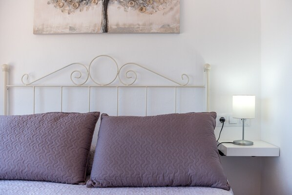 Un lit avec une tête de lit en métal, deux oreillers, une table de chevet avec une lampe et une œuvre d’art accrochée au mur.