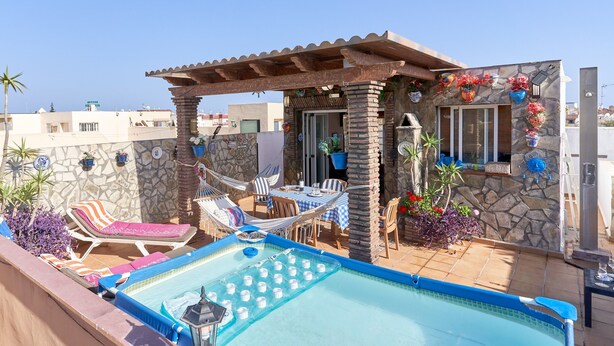 Un área de piscina en la azotea con sillones, una mesa y una pérgola.