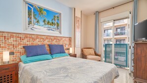 Habitación de hotel con cama, televisión, ventana con vista a la playa y balcón con vistas a otros edificios.